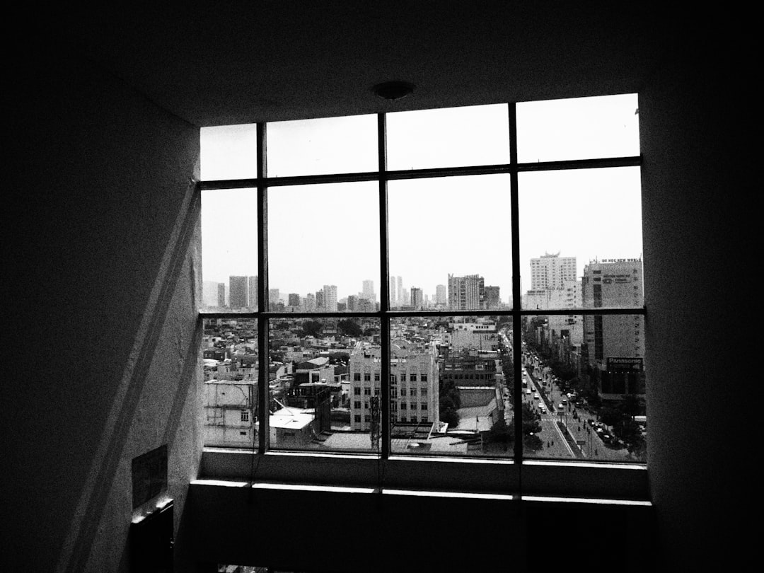 A confident real estate expert stands in a minimalist office, looking out a large window at a city skyline, representing market authority and long-term success.